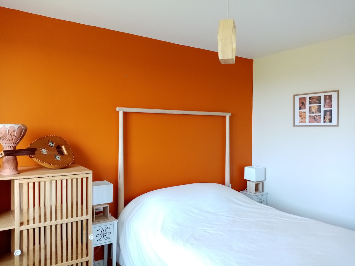 A bright bedroom features an orange accent wall, complementing a spacious bed with a white cover. Adjacent, a wood lattice nightstand holds a decorative item and a lamp. Soft lighting emanates from a hanging pendant above, creating a warm atmosphere.