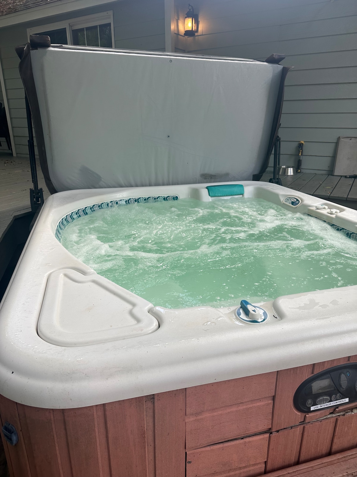 A hot tub is shown with bubbling water, surrounded by a wooden exterior. The cover is partially opened, allowing light to reflect off the surface of the water. The area is framed by a light-colored wall, enhancing the relaxing ambiance.
