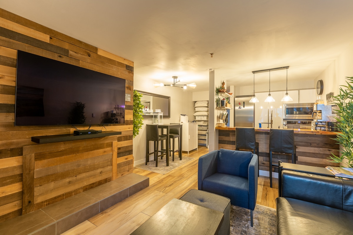 A modern living space features a large wooden accent wall behind a wall-mounted television. Comfortable seating options, including two blue chairs and a sleek black couch, are arranged around a coffee table. The open layout reveals a dining area and a well-equipped kitchen visible in the background.