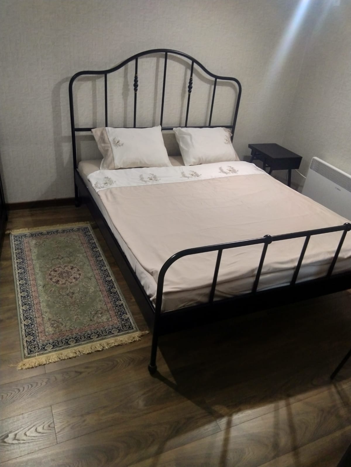 A neatly arranged sleeping area features a black metal bed with soft, light-colored bedding and two decorative pillows. A small nightstand is positioned next to the bed. The hardwood floor is complemented by a rectangular patterned rug placed beneath the bed.