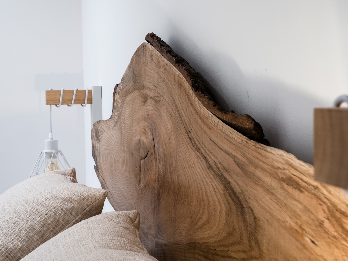 A natural wooden headboard made from a large, curved slab of wood is displayed above two cream-colored pillows. A stylish bedside lamp with a geometric design is positioned to the left, providing a simple yet modern look.