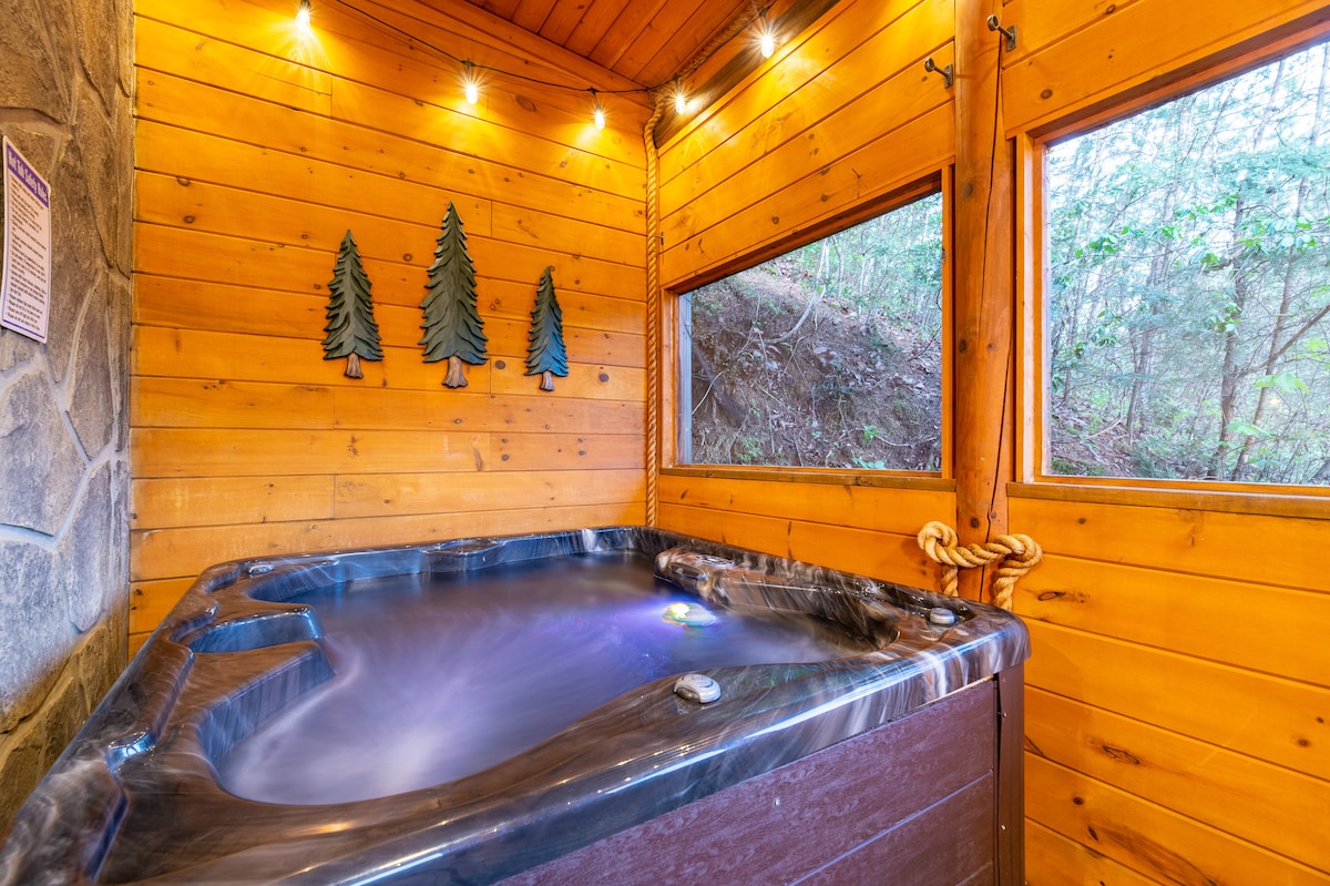 A private hot tub area is surrounded by warm wooden paneling and softly illuminated by recessed lighting. Large windows provide views of the natural surroundings, enhancing the relaxing atmosphere. The hot tub features an inviting water surface, perfect for unwinding after a day of exploration.