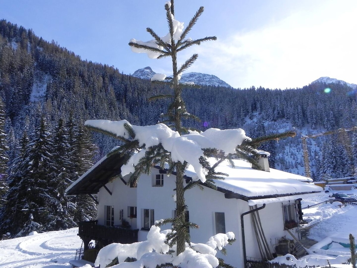 A white chalet is surrounded by snow-covered trees and mountains. The roof is blanketed with snow, while sunlight illuminates the scene, creating a serene winter atmosphere. The lush coniferous forest extends into the background, providing a scenic and peaceful setting.
