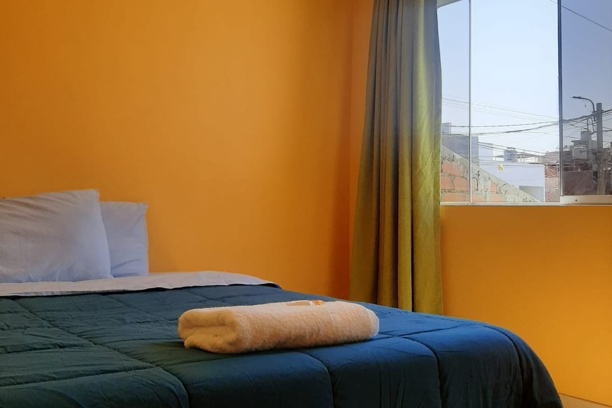 A bright bedroom features an orange wall and a large window with green curtains. A spacious bed is dressed in a dark blue cover, complemented by two white pillows. A neatly rolled towel is positioned on the bed.