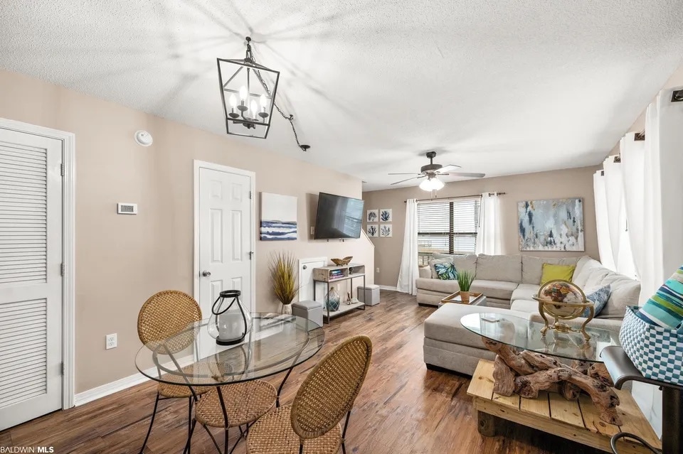A spacious living area is showcased with an L-shaped sofa, a wall-mounted television, and decorative artwork. A glass dining table surrounded by chairs is positioned near the entry. Natural light filters through large windows, highlighting the wood-look flooring and neutral-toned walls.