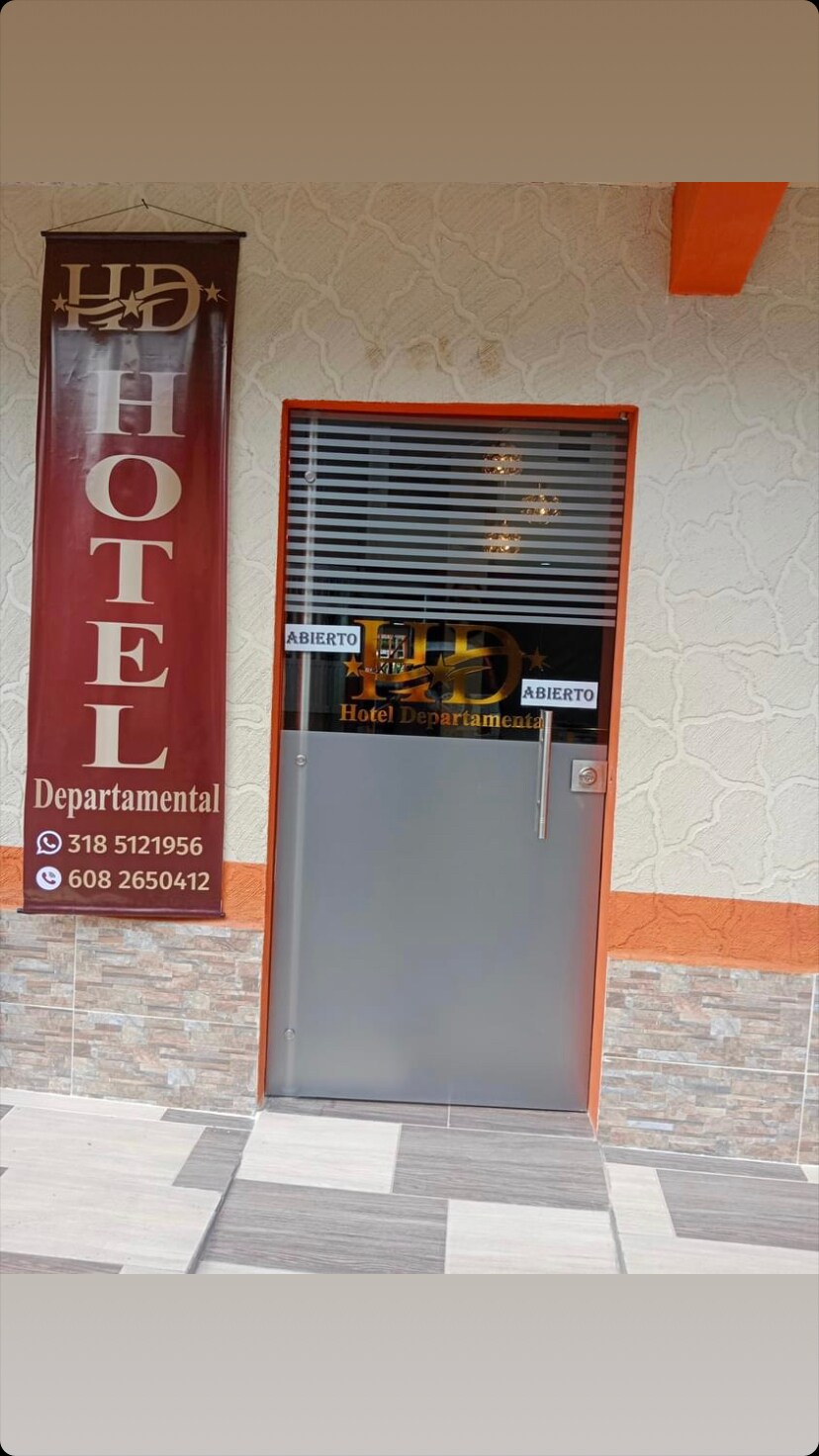 The main entrance of the hotel is shown, featuring a sleek gray door with a simple handle. A vertical banner lists hotel information, including contact numbers. The exterior wall displays a textured finish, complemented by a warm color palette and an overhanging ledge above the entrance.