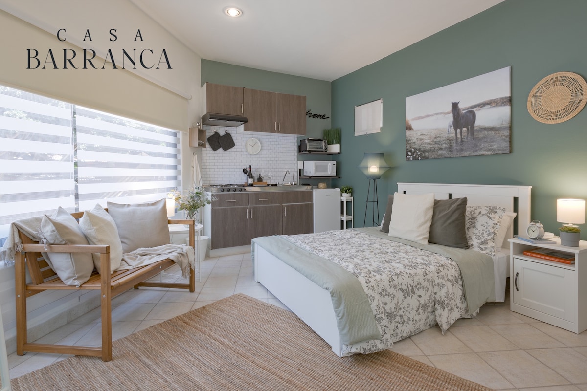 A cozy studio space features a spacious bed dressed in light-colored linens. A kitchen area equipped with essential appliances and a comfortable seating arrangement are visible. Natural light filters through sheer window coverings, while a piece of wall art depicting a horse adds a serene touch.