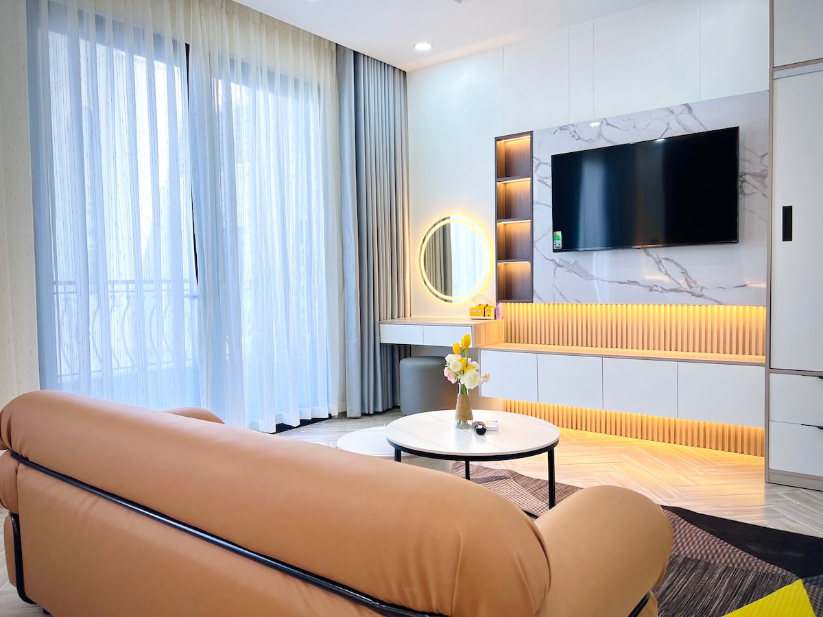 A modern living area features a large, soft sofa in a light tan color. A round coffee table sits in front of it, and a wall-mounted television is positioned above a sleek console with built-in lighting. Natural light filters through sheer curtains, enhancing the spacious feel.