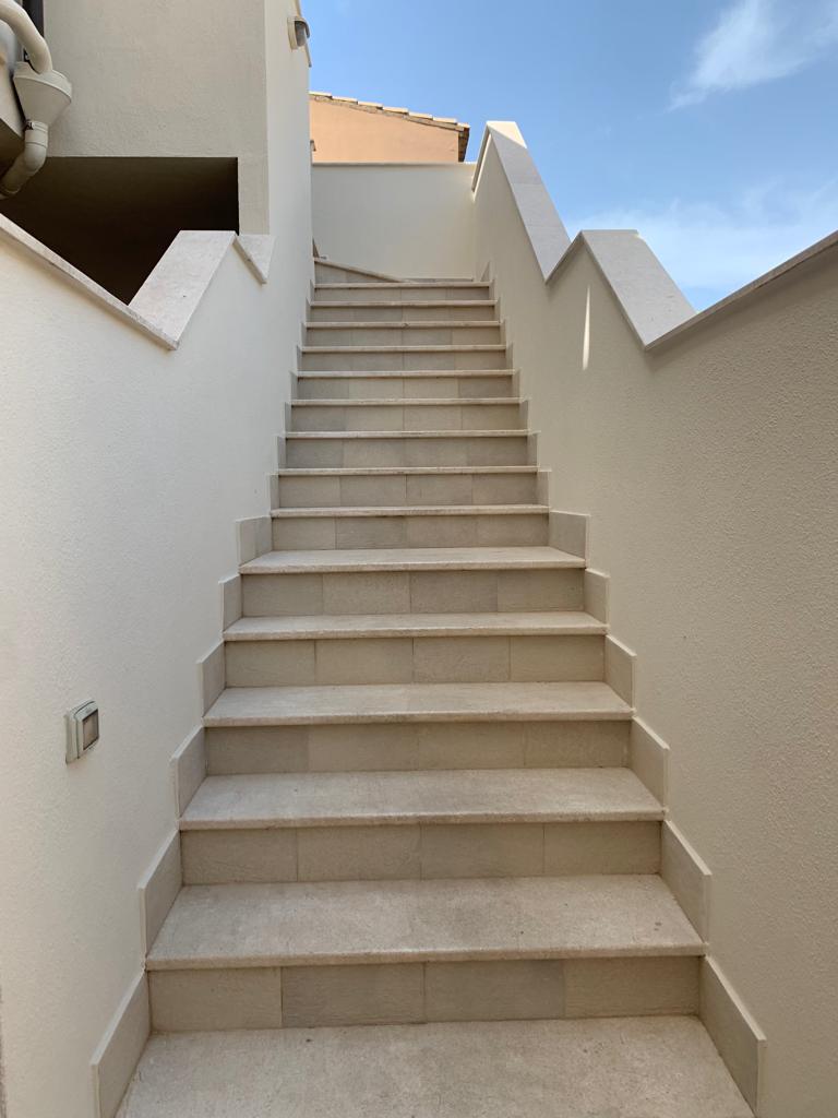 A well-maintained outdoor stairway is depicted, featuring a smooth stone surface and white walls. Natural light illuminates the steps, while the clear blue sky is visible at the top, creating a sense of openness and accessibility.