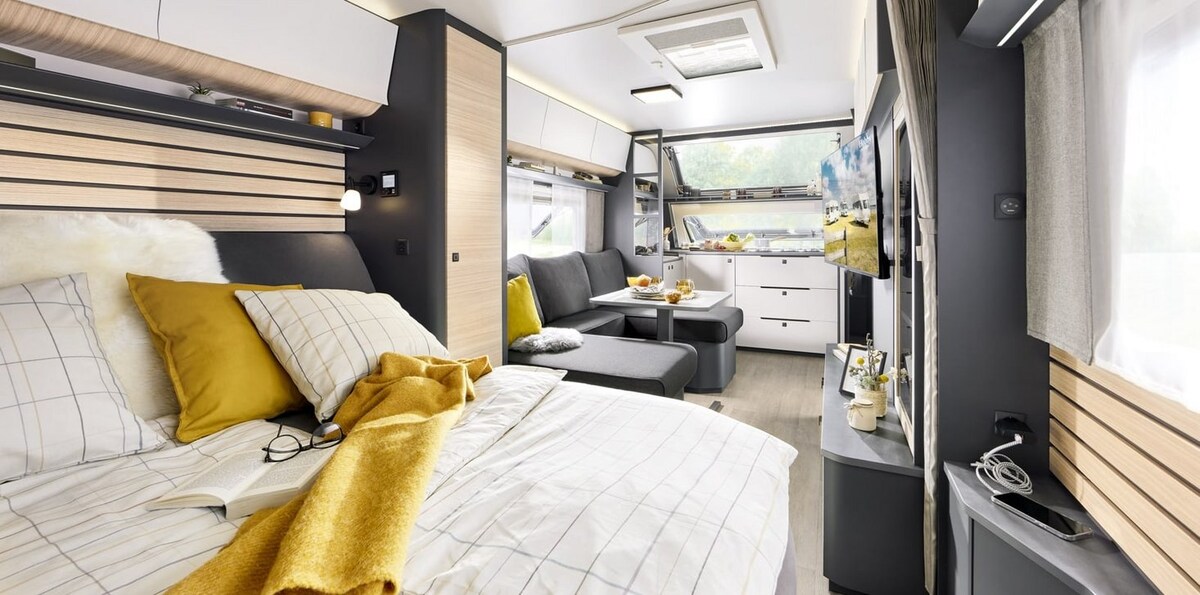 A cozy interior features a spacious bed adorned with soft yellow accents and patterned bedding. A comfortable seating area includes a gray couch and a central table set for breakfast, while large windows bring natural light into the well-designed living space.