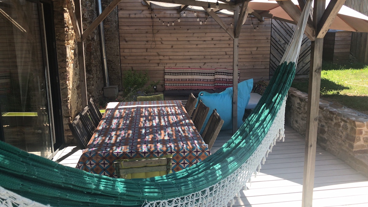 An outdoor seating area is featured, showcasing a large dining table surrounded by several chairs. A hammock is hung nearby, inviting relaxation. The space is adorned with patterned textiles, and a cozy seating option is visible in the background, enhancing the outdoor setting.
