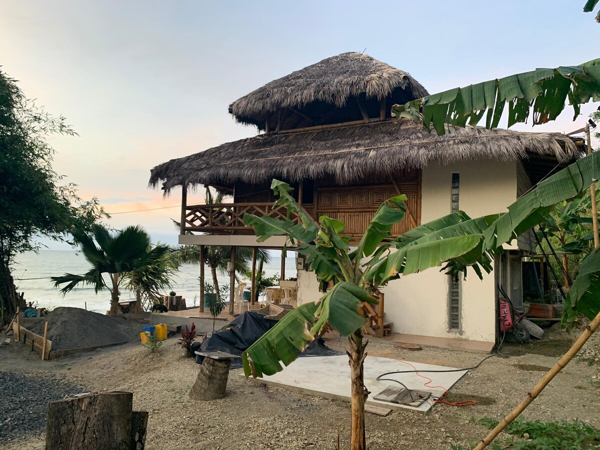 A two-story bamboo house is shown, featuring a thatched roof and wooden accents. The ground level includes a concrete area surrounded by tropical plants. The beach and ocean are visible in the background, accented by a soft sunset light.