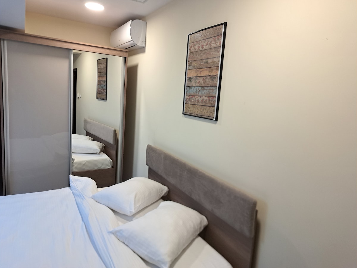 A spacious sleeping area features a large bed with comfortable pillows and a neutral-colored headboard. A large mirror reflects the bed and room decor. A framed artwork hangs on the wall, adding character to the space, while an air conditioning unit is mounted above.