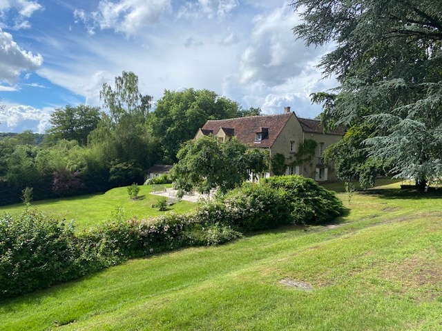 A charming property is set within a lush green landscape, flanked by trees and flowering shrubs. The house features a rustic exterior with a sloped roof and multiple windows, welcoming natural light. The expansive lawn offers an inviting space for relaxation and outdoor activities.