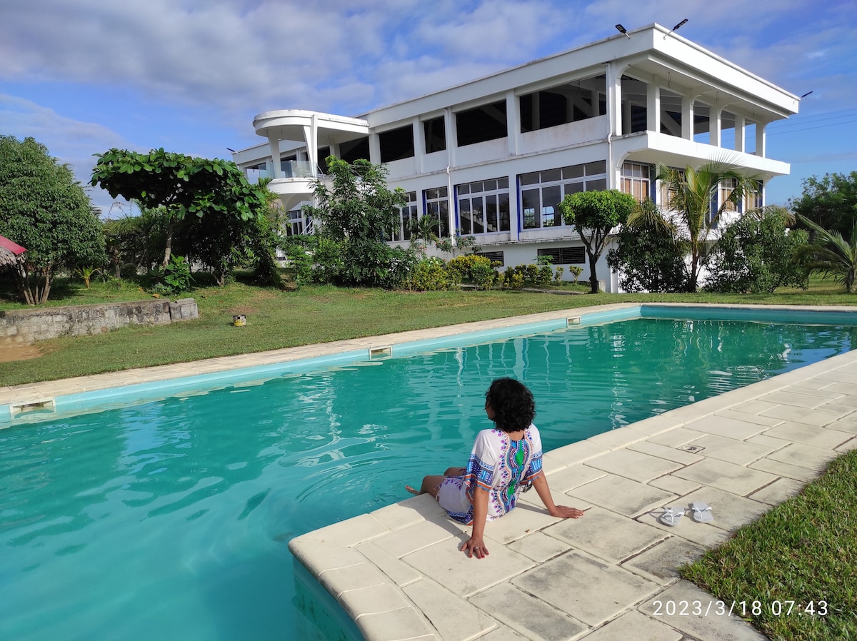 A serene pool area is shown, featuring a long, turquoise swimming pool surrounded by well-maintained grass. A person is seated on the edge, with the spacious white building visible in the background, characterized by large windows and a bright exterior.