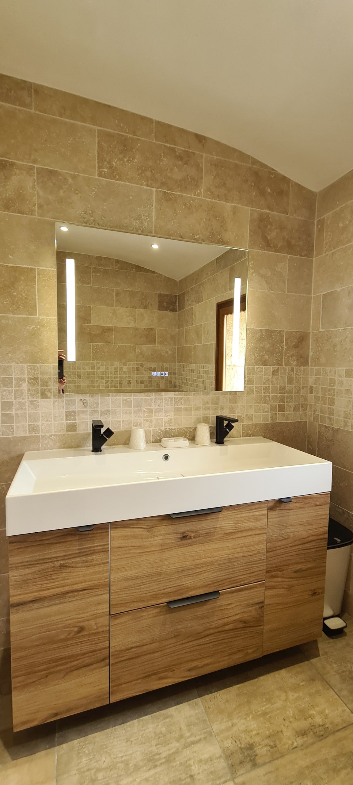A well-lit bathroom features a spacious vanity with a dual sink and wooden cabinetry. Above the sinks, a large mirror reflects the ambient lighting, enhancing the room's openness. Natural stone tiles adorn the walls and floor, providing a warm, neutral backdrop.