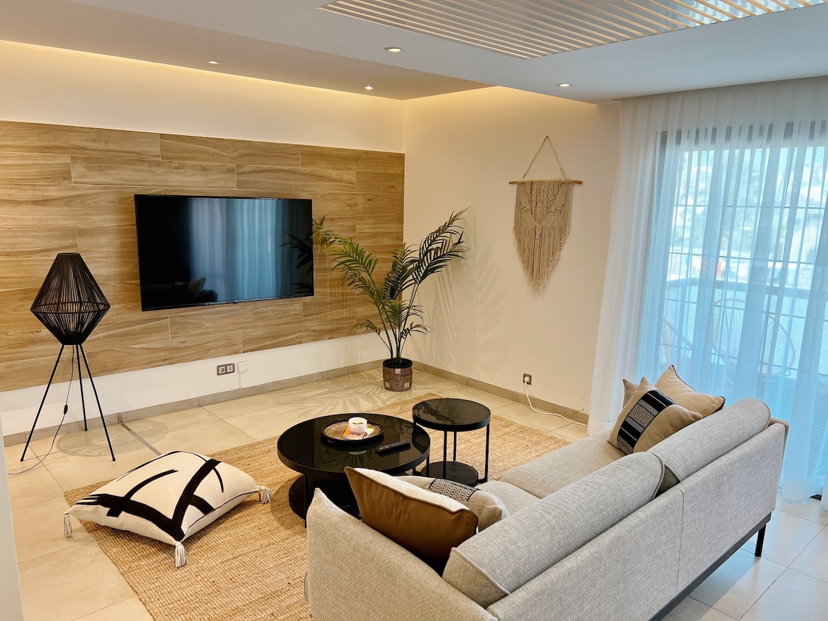 The living area features a comfortable sectional sofa facing a large wall-mounted flat-screen TV. A cozy area rug anchors the space, complemented by a modern coffee table. Natural light filters in through sheer curtains, while a potted plant adds a touch of greenery.