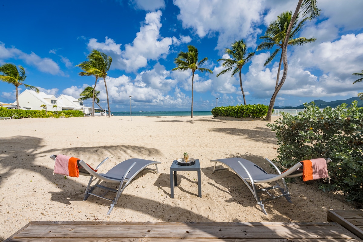 Two beach loungers are positioned on soft sand, with colorful towels draped over the arms. A small table sits between them, adorned with a potted plant. Lush palm trees sway gently in the breeze, and a view of the calm sea and sky enhances the serene atmosphere.