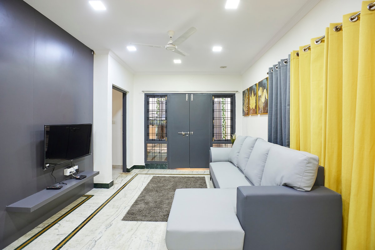 A comfortable living area features a gray sofa positioned against a dark accent wall. A smart TV is mounted on a sleek shelf, while light gray curtains complement the bright yellow drapes. A soft area rug is placed on the marbled floor, enhancing the cozy atmosphere.