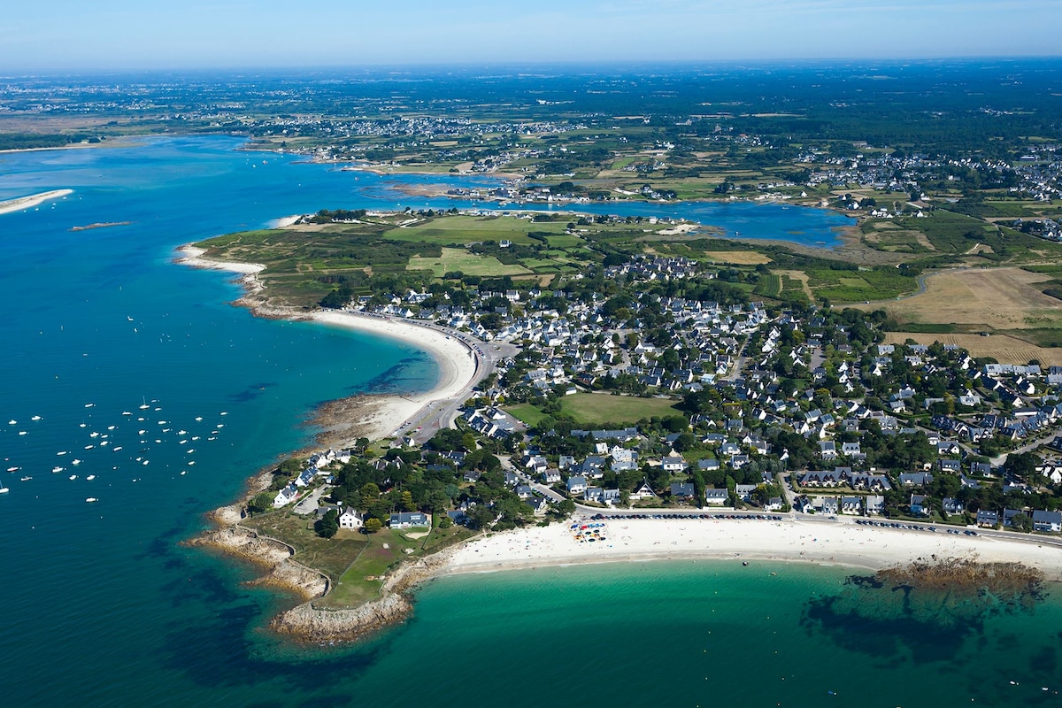 An aerial view showcases the coastline lined with sandy beaches, framed by clear turquoise waters. Small boats are scattered along the bay, while a charming village with white buildings and green landscapes is visible in the background, accentuating the serene seaside environment.