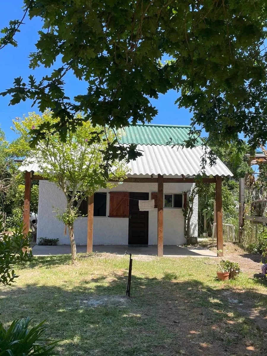 A two-story house is presented, featuring a sloped roof and wooden posts supporting a covered porch. Surrounding greenery enhances the setting, with a lawn area visible. The entrance features a wooden door, leading to a welcoming living space inside.