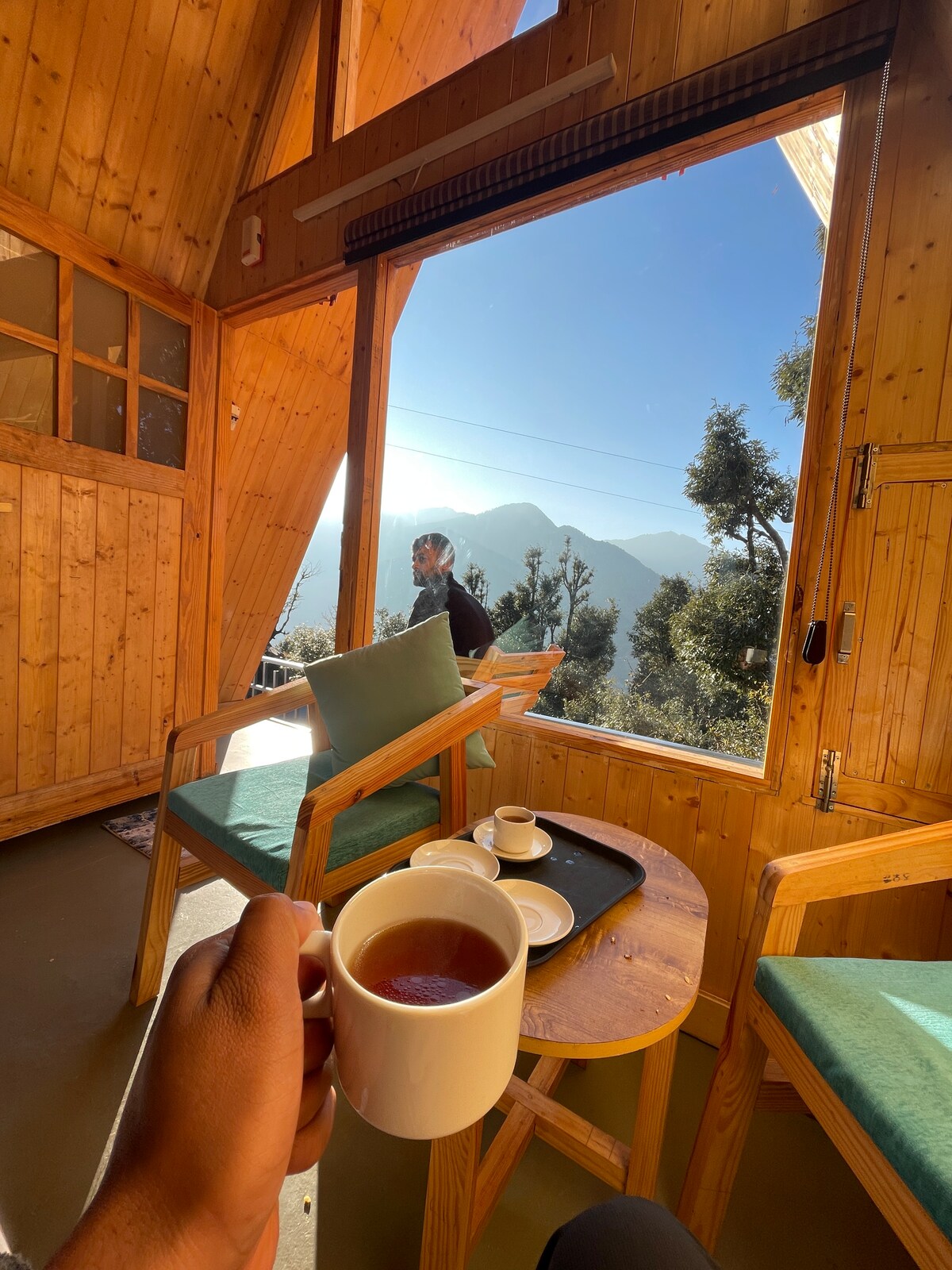 A cozy cabin interior features wooden walls and large windows that frame a breathtaking mountain view. A small table holds a cup of tea and a notepad, while a comfortable chair invites relaxation. Natural light enhances the warm ambiance of the space.