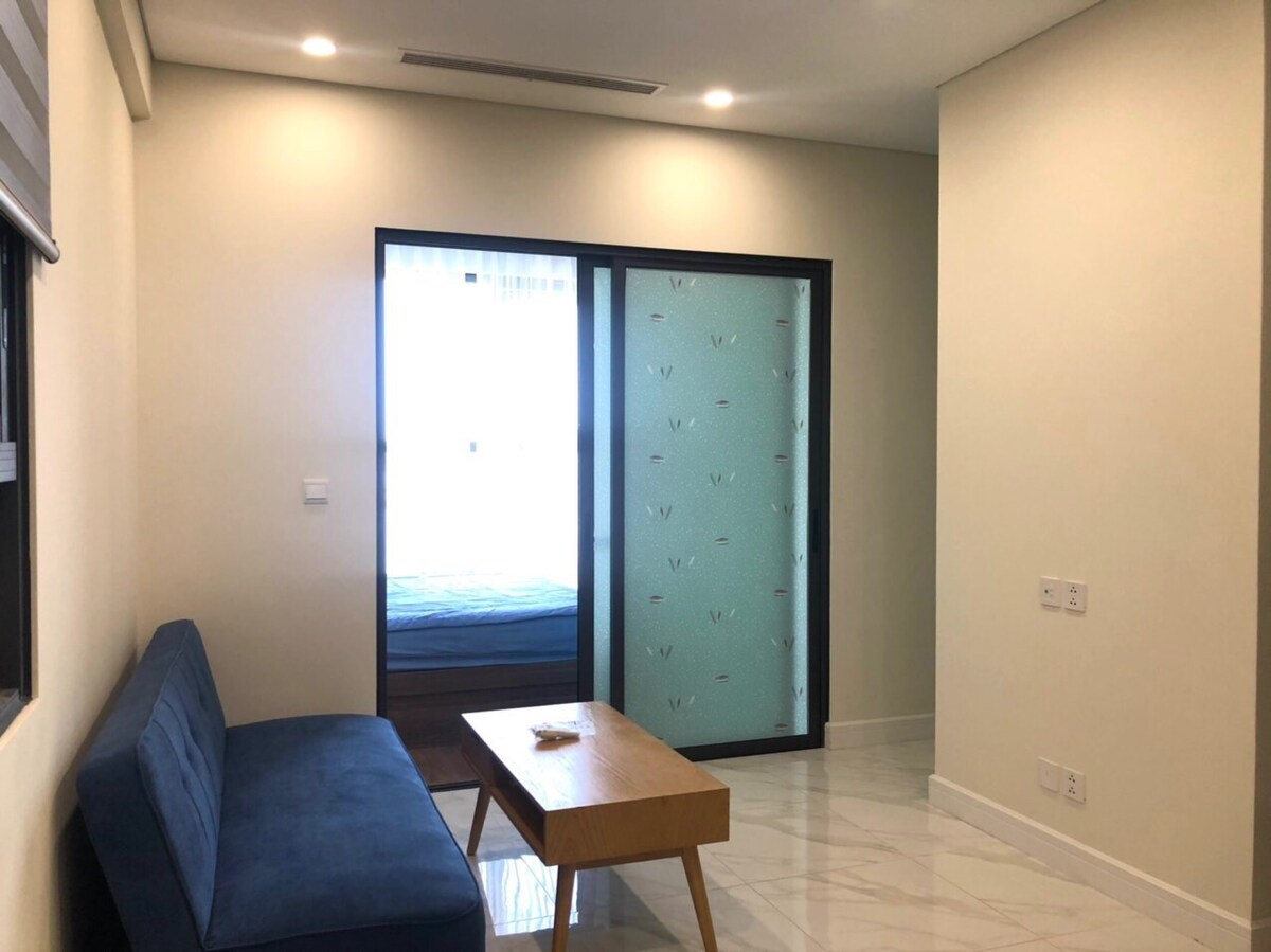 A living area is presented with a navy blue sofa facing a wooden coffee table. A sliding glass door reveals a sleeping area beyond, while natural light filters in, illuminating the white walls and polished floor. The overall design maintains a sleek and modern appearance.