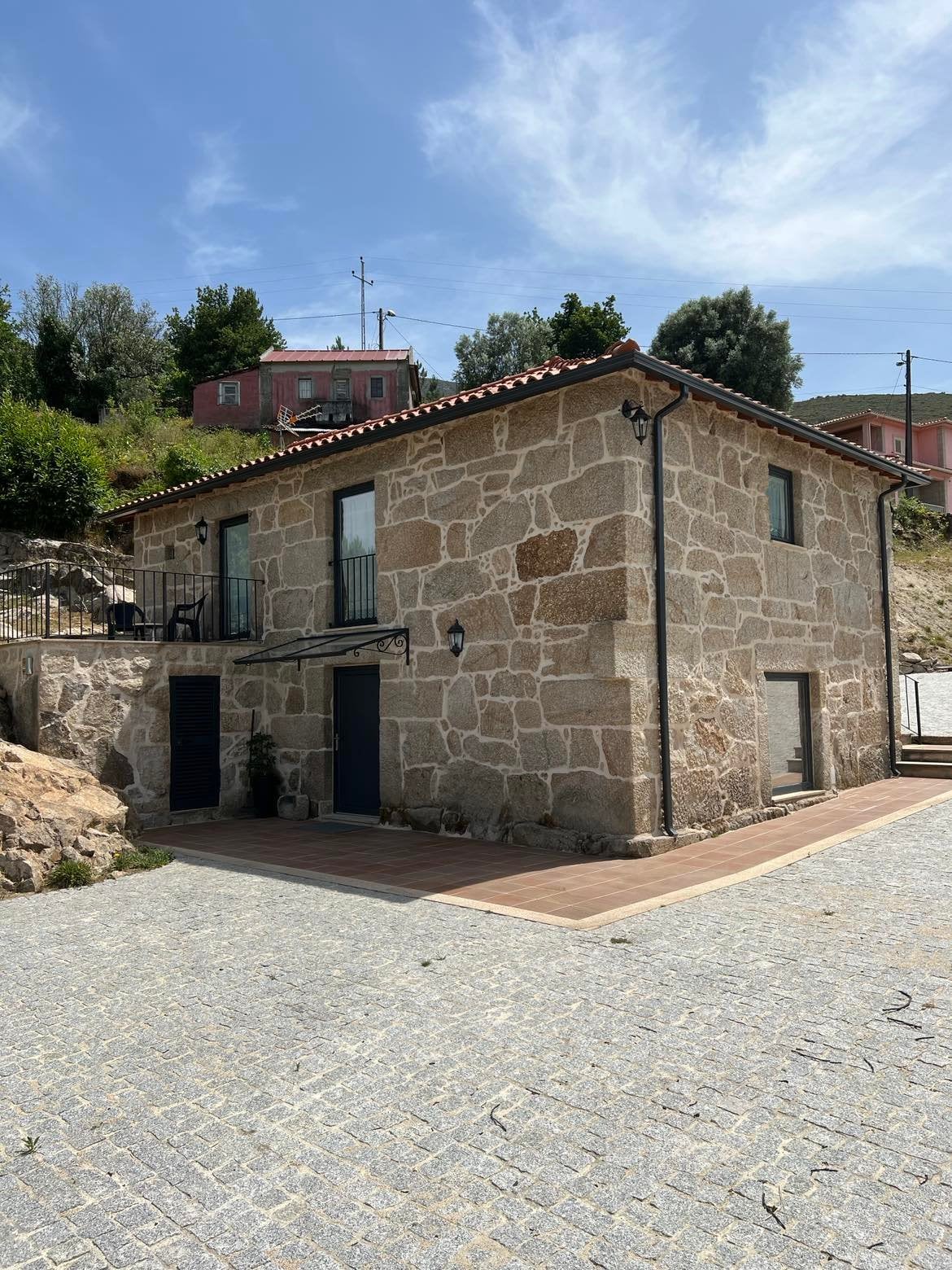 A charming two-story stone house is set against a backdrop of greenery. The exterior features a traditional tiled roof and several windows, allowing natural light to enter. A wooden deck area is visible, surrounded by a gravel pathway and natural landscape.