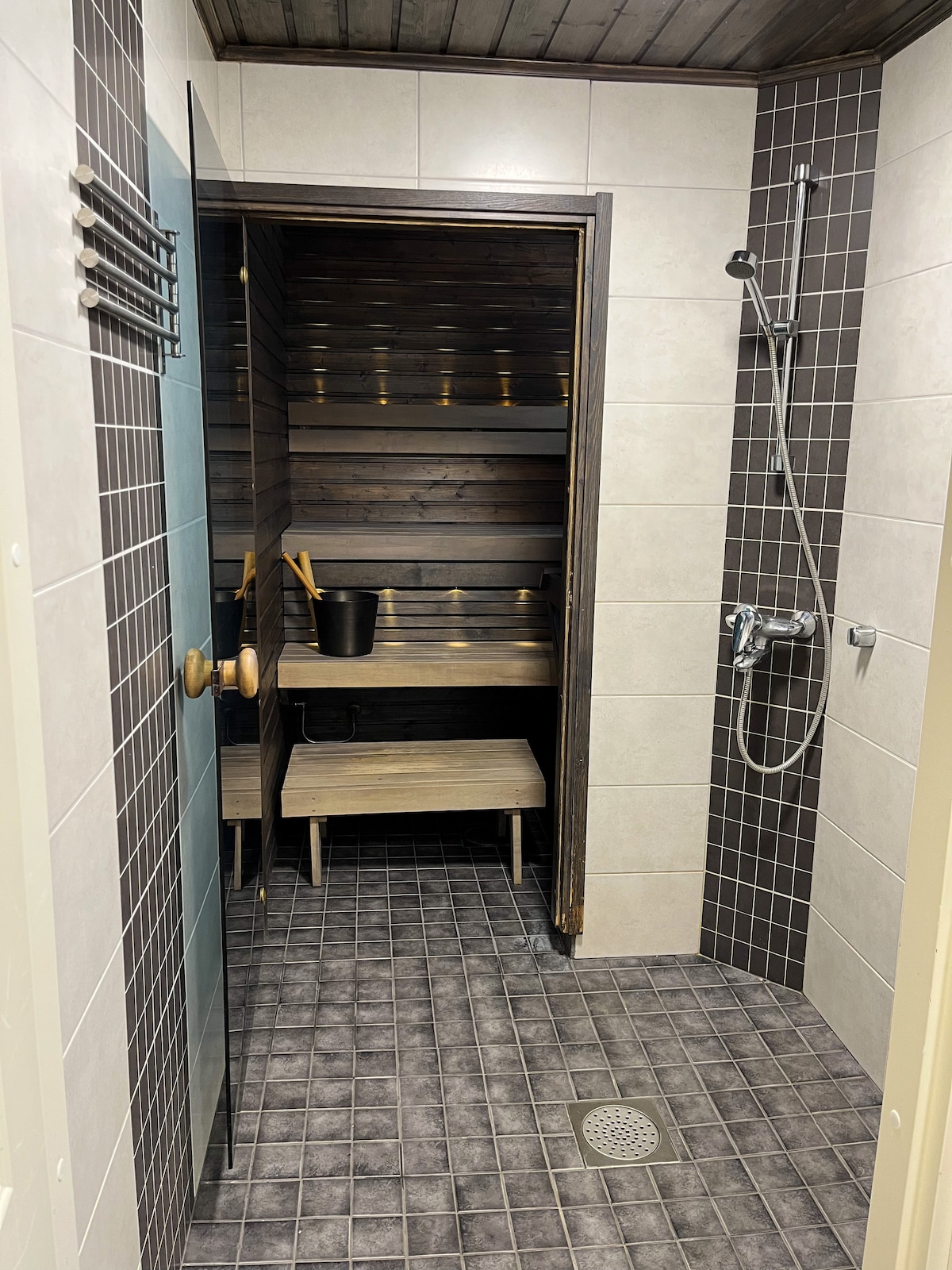 A modern sauna space features wooden seating and a minimalist design. Soft lighting enhances the natural wood tones. A shower area is adjacent, equipped with a showerhead and tiled surfaces. The floor is covered with dark, textured tiles, providing contrast with the lighter wall tiles.