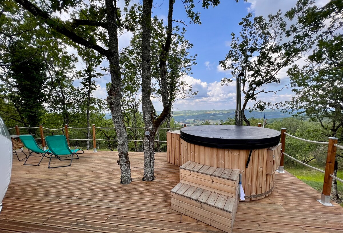 A wooden terrace features a round, wooden hot tub surrounded by trees, offering expansive views of the valley. Two green lounge chairs are positioned nearby, inviting relaxation under the open sky. The deck's natural aesthetic complements the tranquil surroundings.