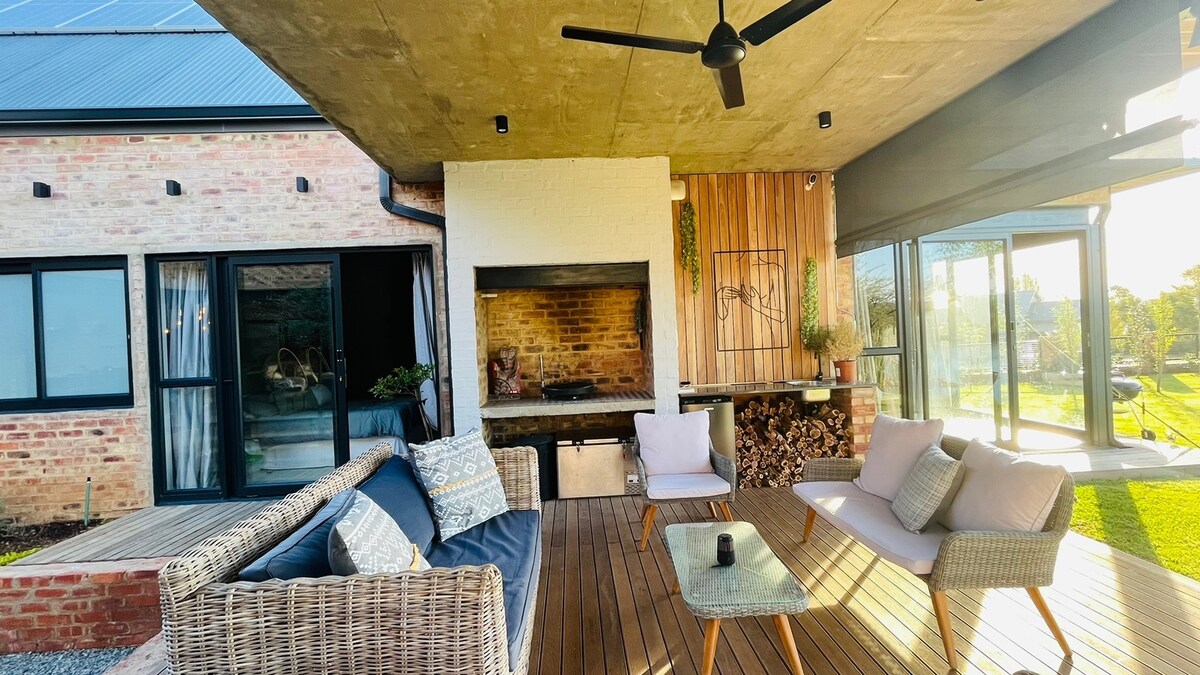 An outdoor seating area is shown with a comfortable sofa and chairs arranged around a wooden table. A built-in barbecue is present, with stacked firewood nearby. Large glass doors open to the interior, allowing natural light to flow in, complemented by a ceiling fan overhead.