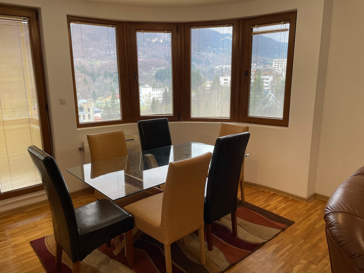 A dining area features a glass table surrounded by four chairs, two upholstered in black and two in tan. Large windows curve around the space, offering views of the mountains and nature, with ample natural light illuminating the wooden flooring.
