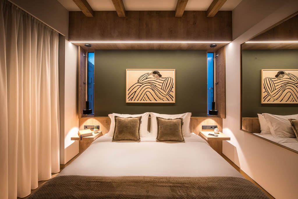 A cozy bedroom is featured with a spacious bed, complemented by soft pillows and two bedside tables. The warm lighting is framed by wooden beams, and a large mirror enhances the room's brightness. A piece of artwork is displayed on the wall above the bed.