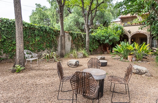 A shared outdoor area is set with stone pathways and seating arrangements. A round wooden table is surrounded by four metal chairs, with various plants and trees adding greenery to the space. Rocky elements and a stone wall create a natural ambience.