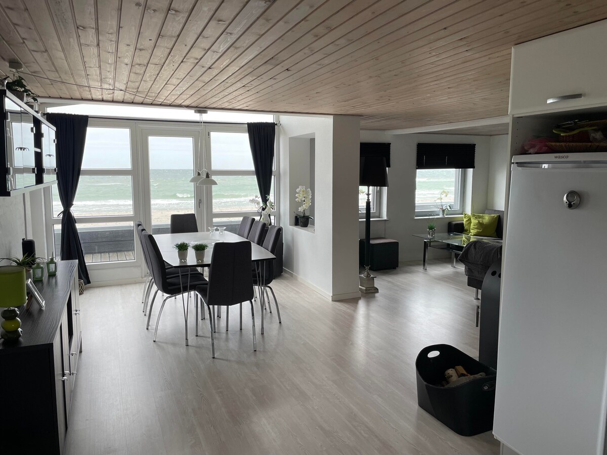 A bright and airy open space features a dining area with a large table and six chairs. Large windows offer views of the sea, while comfortable seating options are arranged around the room. The wooden ceiling adds warmth to the contemporary decor.