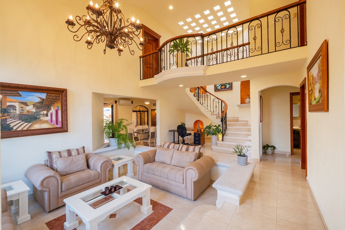 An open living area features two light-colored sofas and a glass coffee table atop a patterned rug. A staircase leads to an upper level, framed with a decorative railing. Natural light floods the space through large windows, accented by decorative plants and wall art.