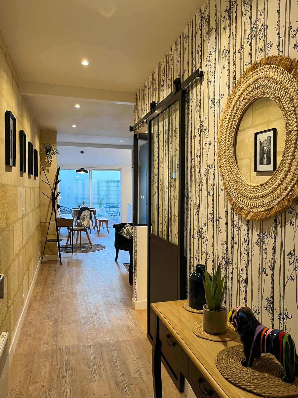 A well-lit hallway features patterned wallpaper and wooden flooring. Framed pictures are displayed along the wall. A decorative mirror hangs above a console table adorned with plants and small decorative items. At the end of the hallway, an open area with dining chairs and a table is visible.