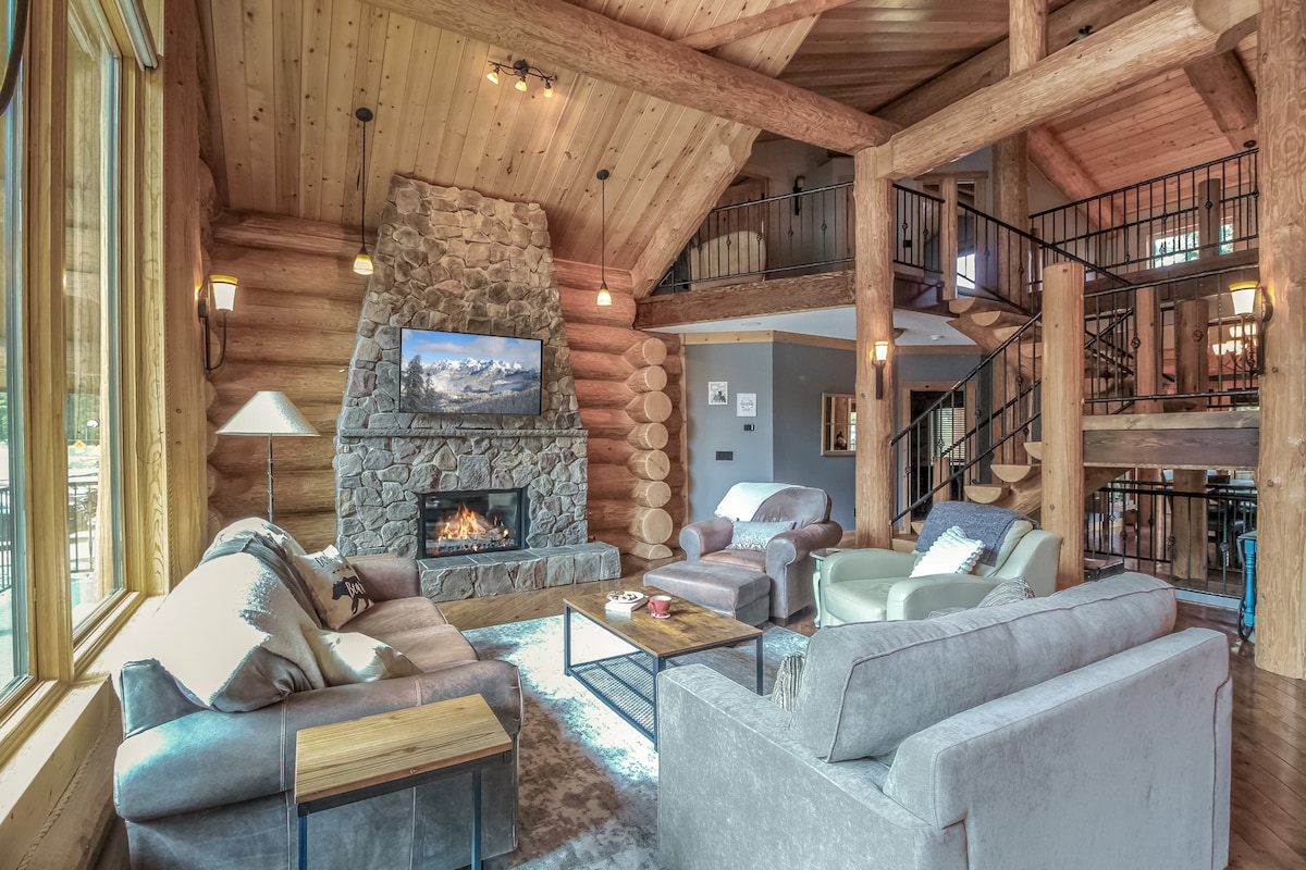 The spacious living area features large log beams and a stone fireplace, providing a focal point under vaulted ceilings. Comfortable seating options include two sofas and an armchair, arranged around a coffee table. Large windows offer views of the surrounding nature, allowing natural light to fill the space.
