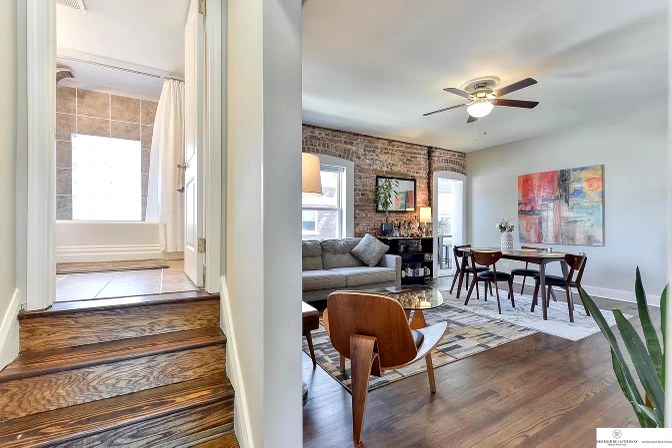 A glimpse into the open living area showcases a comfortable sofa and a wooden dining table surrounded by chairs. Natural light floods through the windows, illuminating the stylish decor, including a colorful artwork on the wall. The entry to a bathroom is visible in the background.