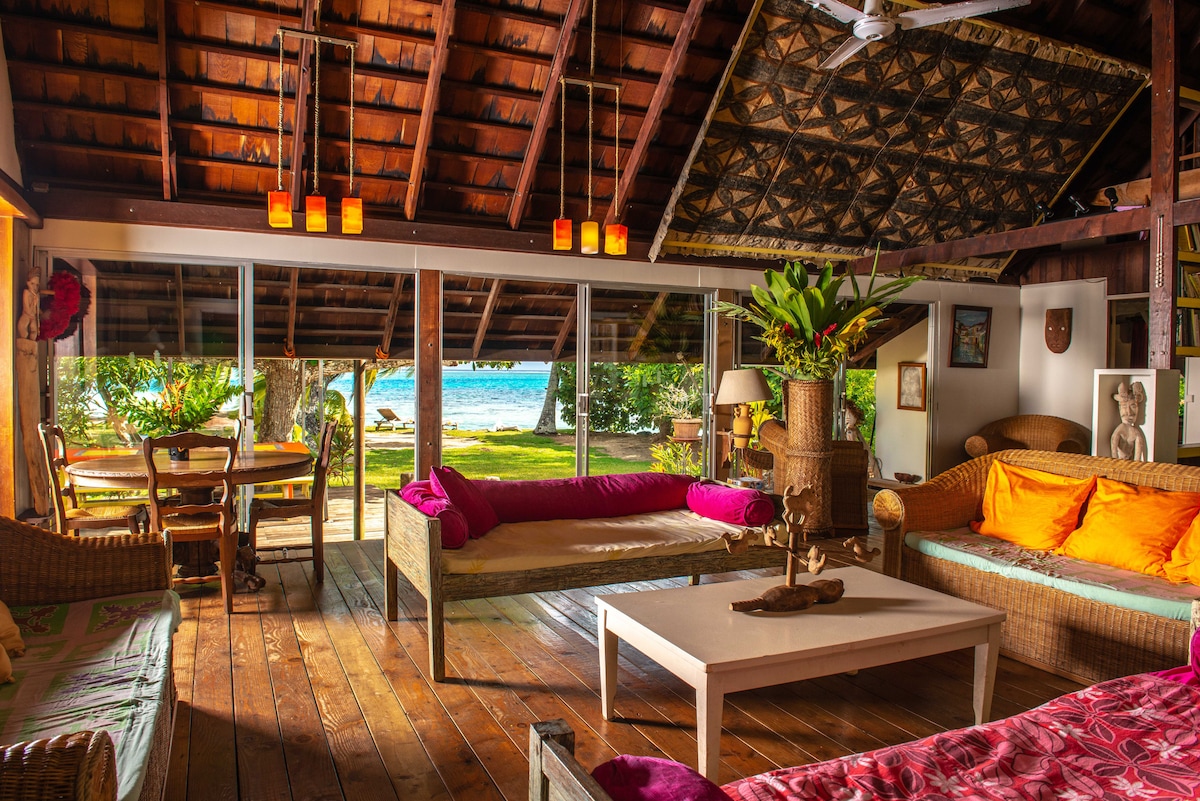 The spacious living area features large glass doors opening to a view of the sea, allowing natural light to fill the room. Rattan furniture, including a coffee table and sofas adorned with colorful cushions, create a welcoming environment. A dining table is placed nearby, surrounded by lush greenery.