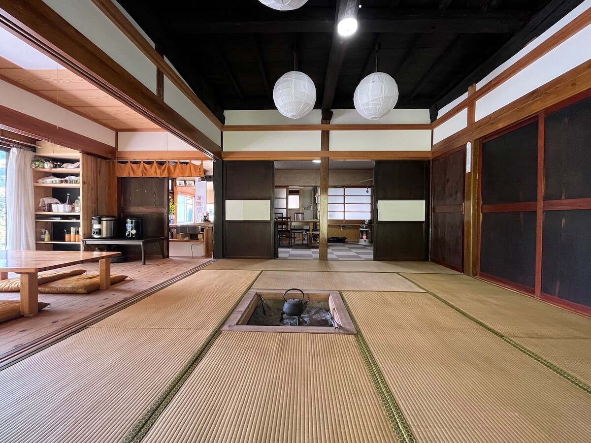 A traditional Japanese room features tatami flooring and shoji screens, with a sunken hearth at the center. Two lantern-style ceiling lights provide soft illumination. Open spaces connect to a modern kitchen area, creating an inviting layout for gatherings.