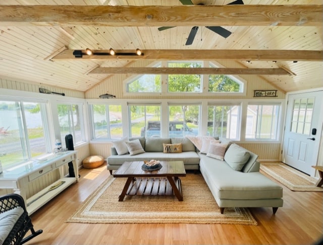 A bright sunroom features high, vaulted ceilings with wooden beams and large windows that allow ample natural light. Comfortable seating is arranged around a central coffee table, complemented by a decorative rug. Fresh greenery is visible outside, enhancing the inviting atmosphere.