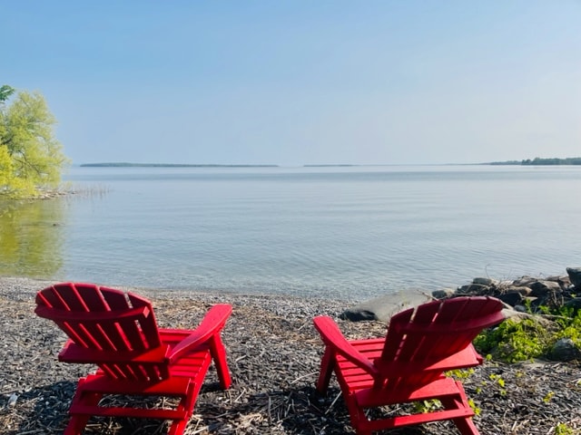 Two red Adirondack chairs are positioned on a pebbled beach, facing a calm, expansive lake. The tranquil water reflects the clear blue sky, while greenery is visible in the background, creating a serene outdoor setting.