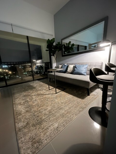 A minimalist living area features a futon sofa adorned with decorative pillows. A glass table stands next to the sofa, complemented by two sleek chairs. Large windows provide a view of the exterior, while soft lighting enhances the space's inviting ambiance.