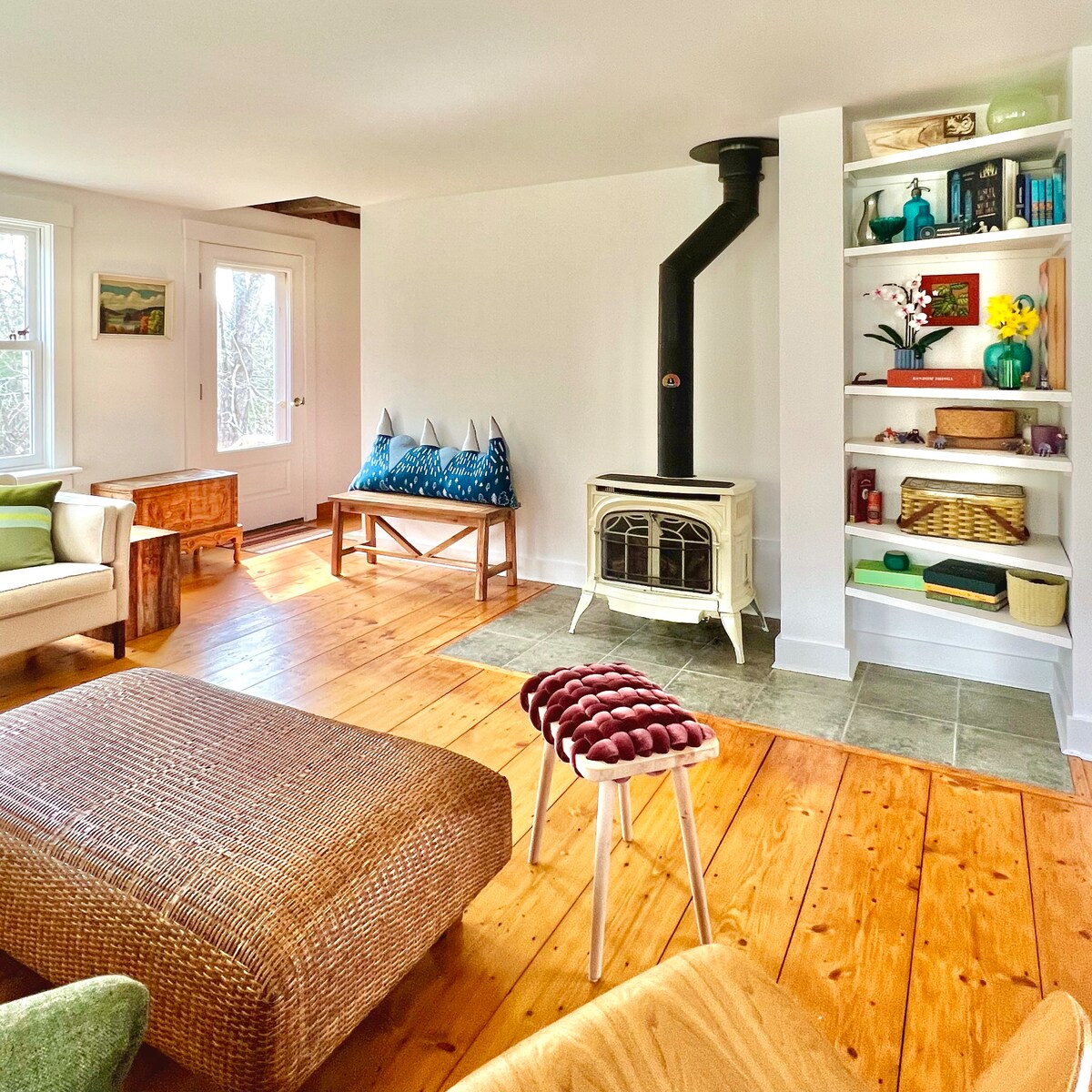 A cozy living area is highlighted by warm wooden flooring and a decorative wood stove. A comfortable chair and a woven pouf offer seating options, while built-in shelves display colorful decor and storage baskets. Natural light streams in through large windows, enhancing the inviting ambiance.