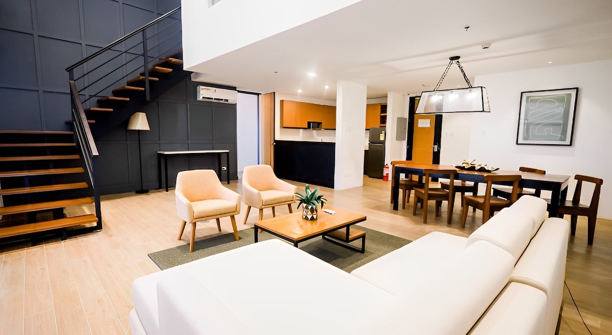 An open living space is showcased, featuring a cozy white sofa, two beige armchairs, and a wooden coffee table adorned with a plant. A dining area with a wooden table and chairs adds to the ambiance, beneath a stylish light fixture. A staircase leads to the upper level.