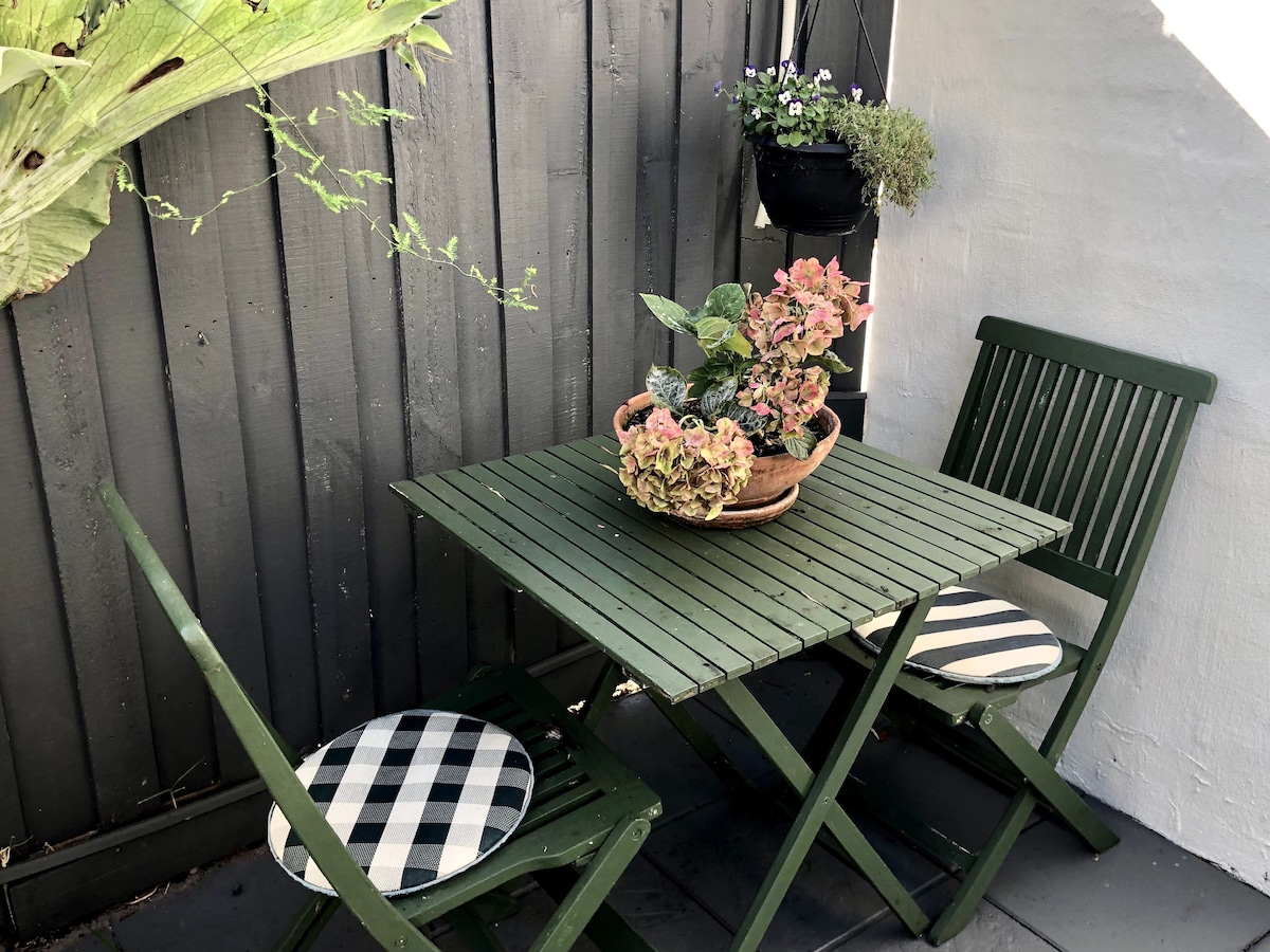 A small outdoor seating area is presented, featuring a green wooden table with a floral centerpiece. Two matching folding chairs with checkered cushions flank the table. A vertical garden can be seen in the background, adding a touch of greenery to the space.