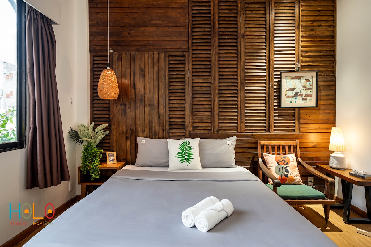 A spacious bedroom is highlighted by wooden wall panels and soft lighting. A large bed adorned with fresh white towels offers comfort. A wooden chair and a small side table complete the inviting space, while a plant and artworks add subtle touches of decor.
