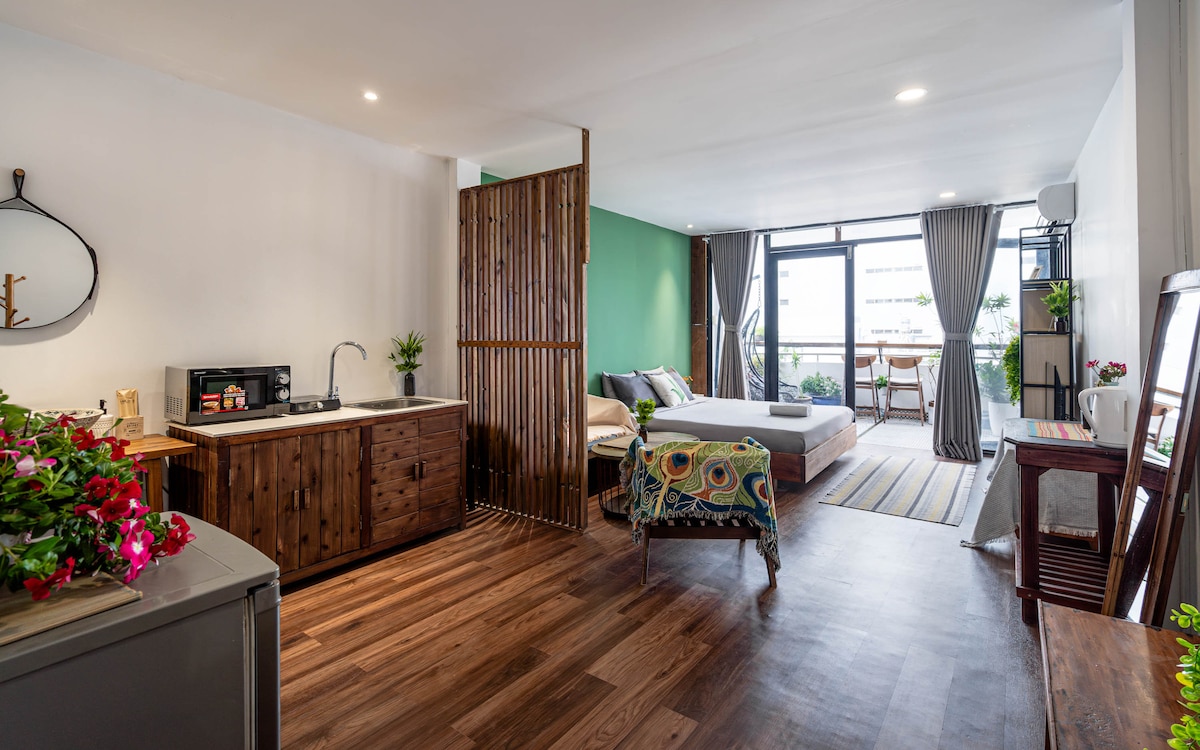 A stylish studio apartment features a cozy sleeping area with a spacious bed and a small kitchenette equipped with a sink and microwave. Natural light fills the space through large windows, complemented by greenery and decorative elements that enhance the inviting ambiance.
