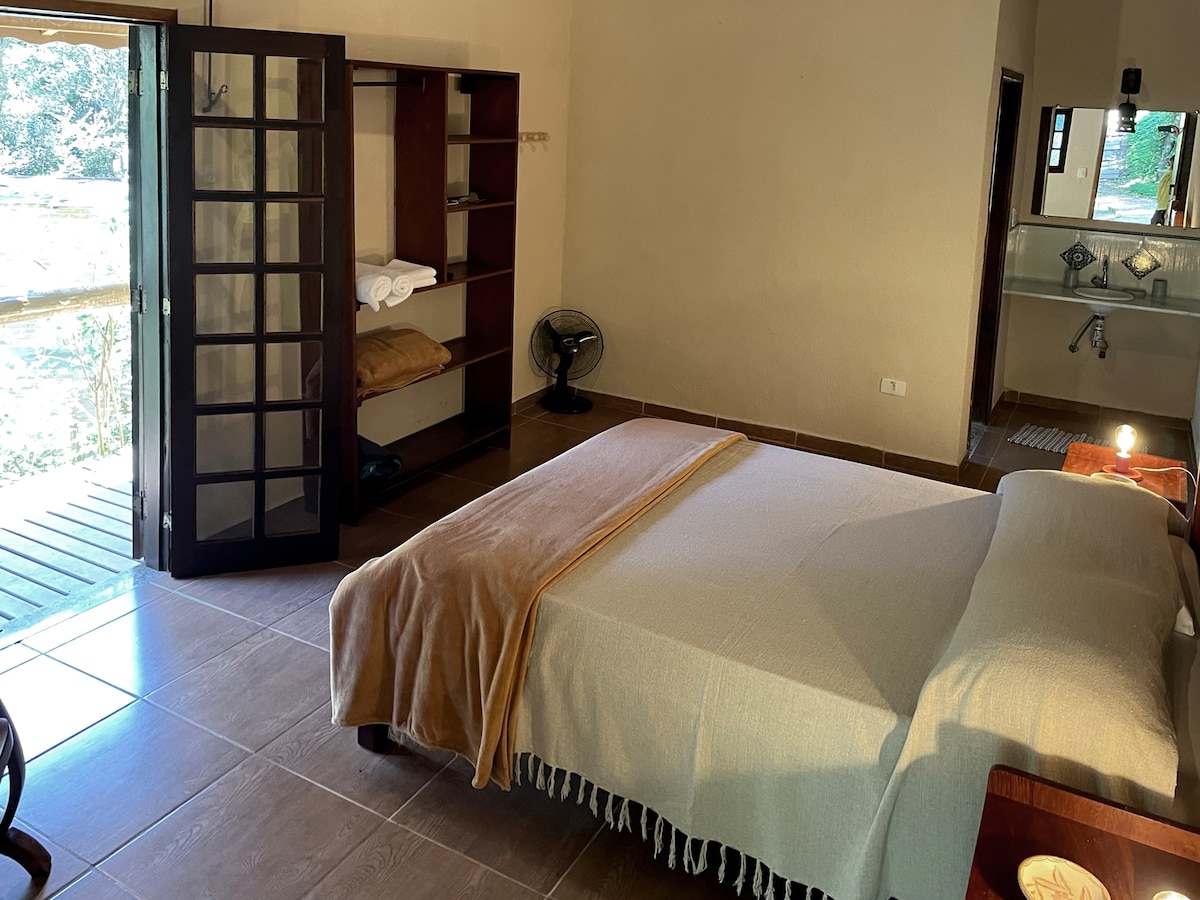 A spacious bedroom is illuminated by natural light from a large open door leading to a wooden deck. The room includes a comfortable bed layered with a neutral-colored blanket, simple wooden furniture, and a fan positioned nearby. A small area with shelves is visible, holding additional linens.
